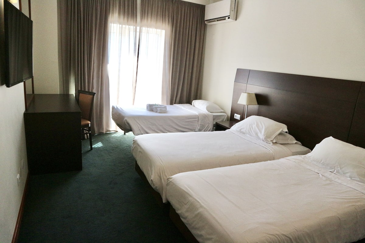 A hotel room with two single beds, a desk, a chair, a TV, and a window with curtains.