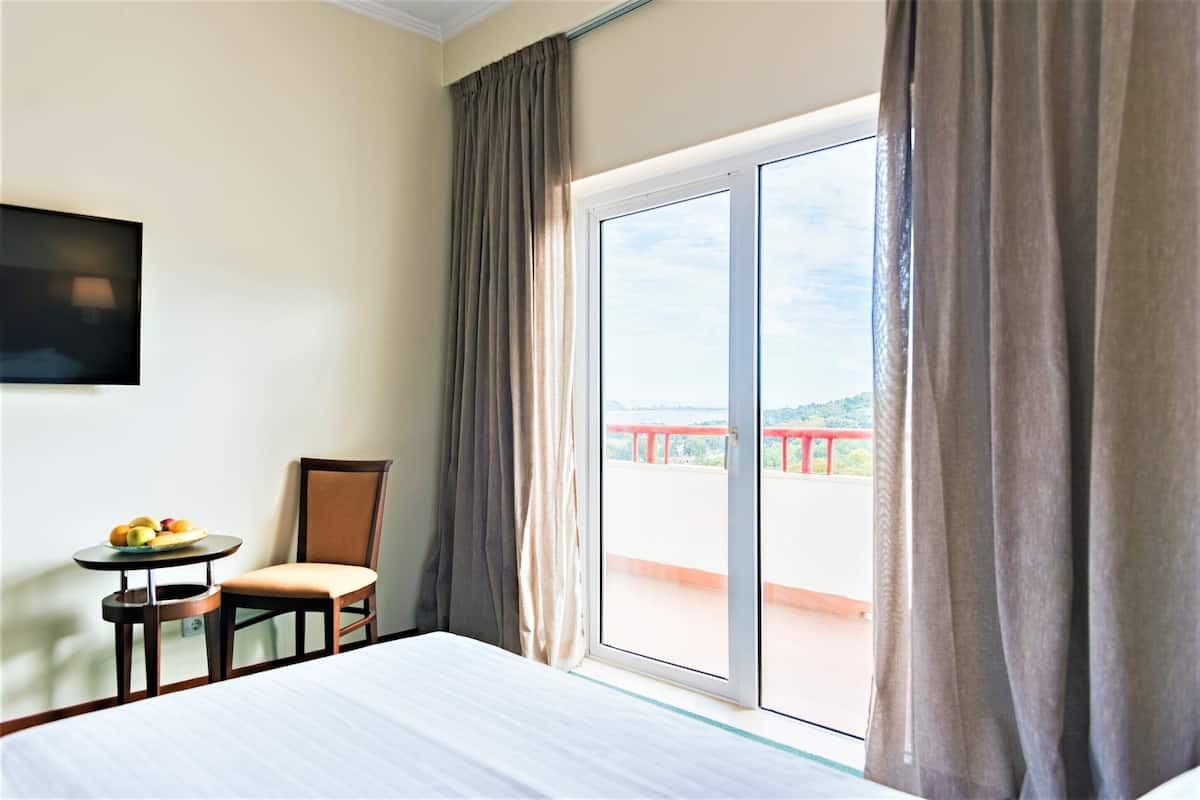 A hotel room with a bed, a chair, a small table with fruit, and a view of a balcony with a railing.