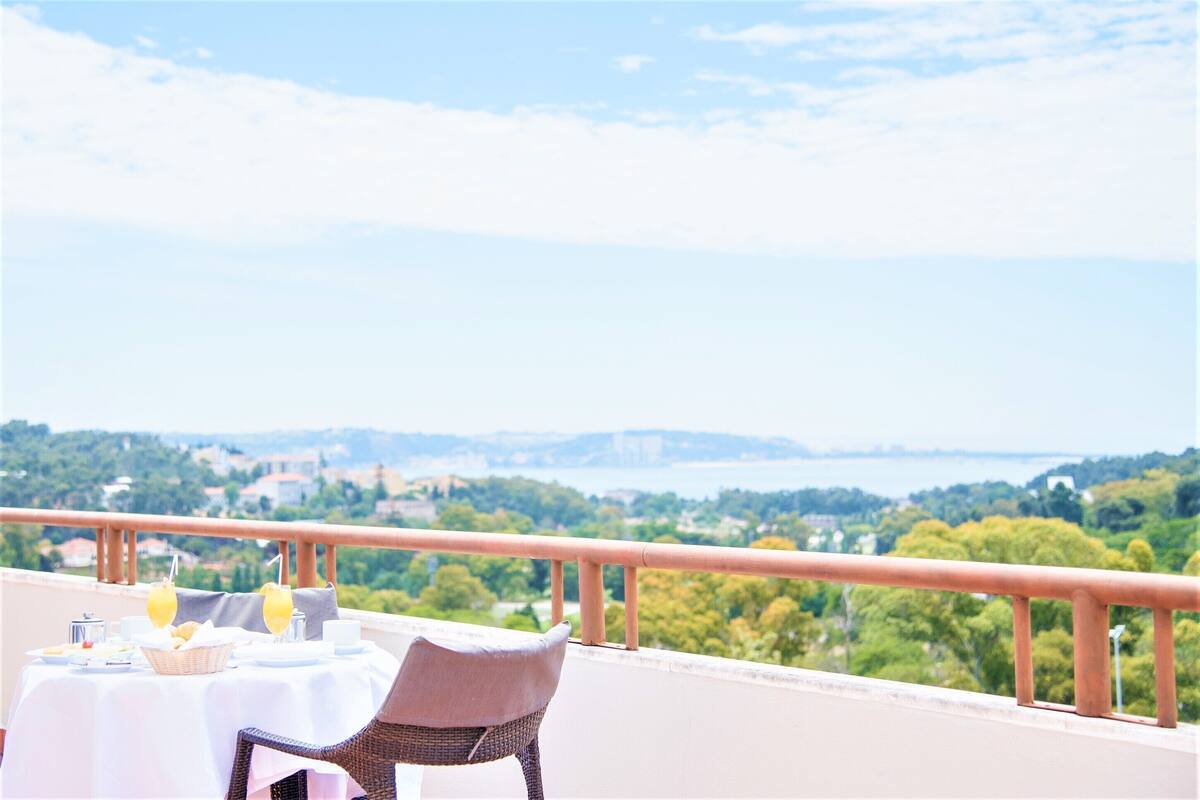 A table set with breakfast items and a wicker chair on a balcony with a view of a green landscape and a body of water.