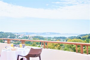 Una mesa dispuesta con alimentos para el desayuno y una silla de mimbre en un balcón con vistas a un paisaje verde y una masa de agua.
