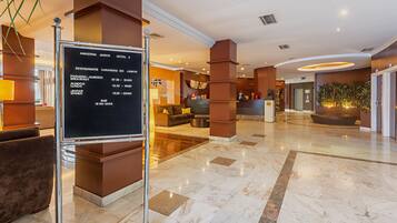 A hotel lobby with a reception desk, a clock, a sofa, and a signboard listing restaurant hours.