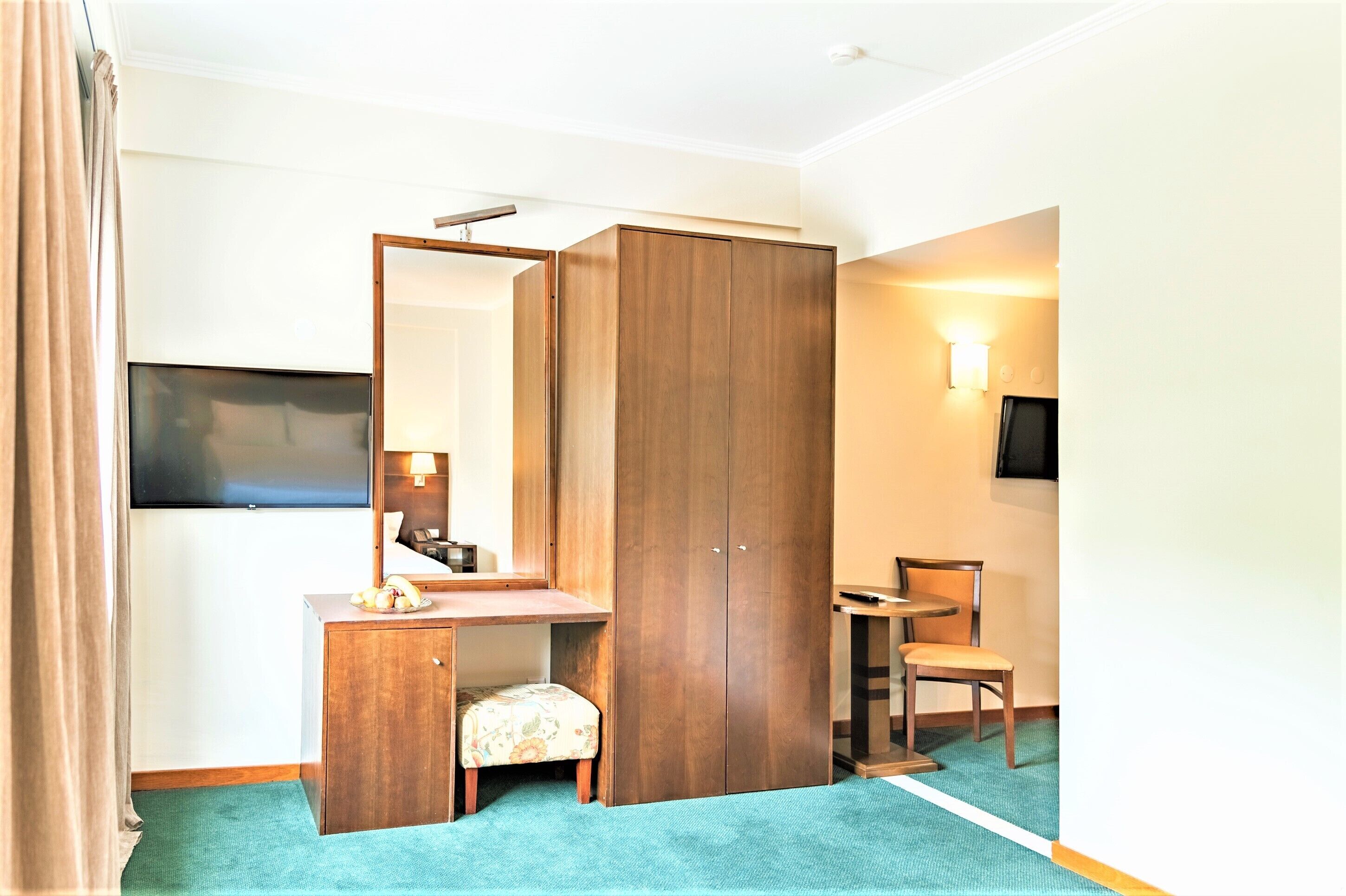 A hotel room with a wooden wardrobe, a desk with a chair, a small table, a television, and a wall-mounted light.