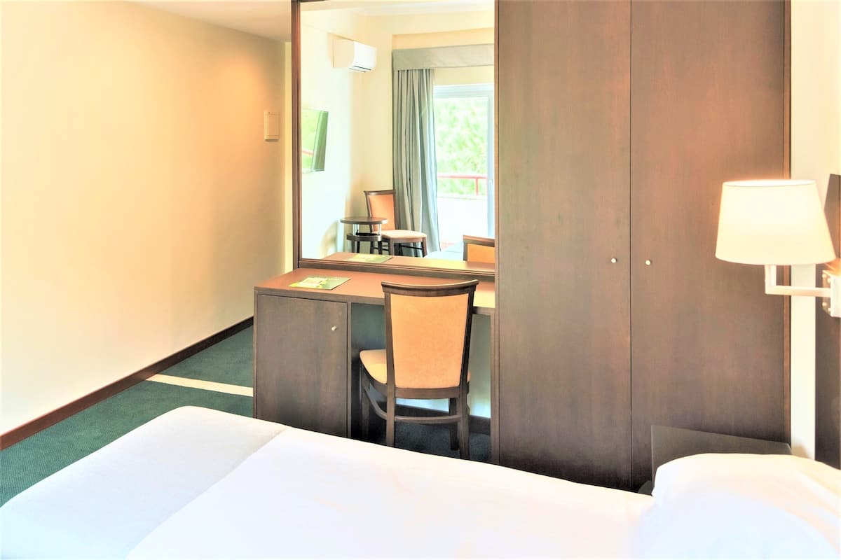 A hotel room with a bed, a desk with a chair, a mirror, and a window with curtains.
