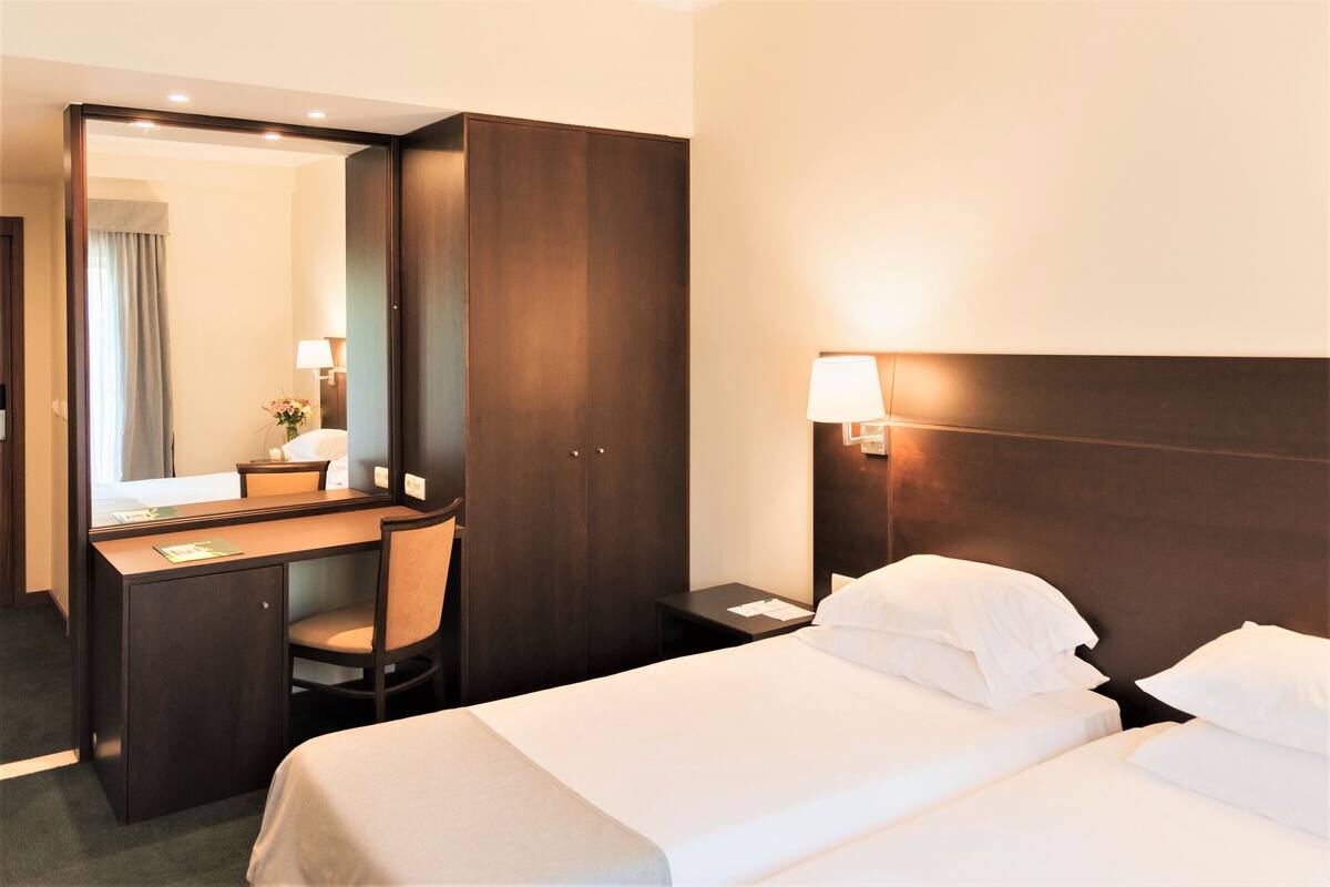 A hotel room with a bed, a desk, a chair, a wardrobe, and a nightstand with a lamp.