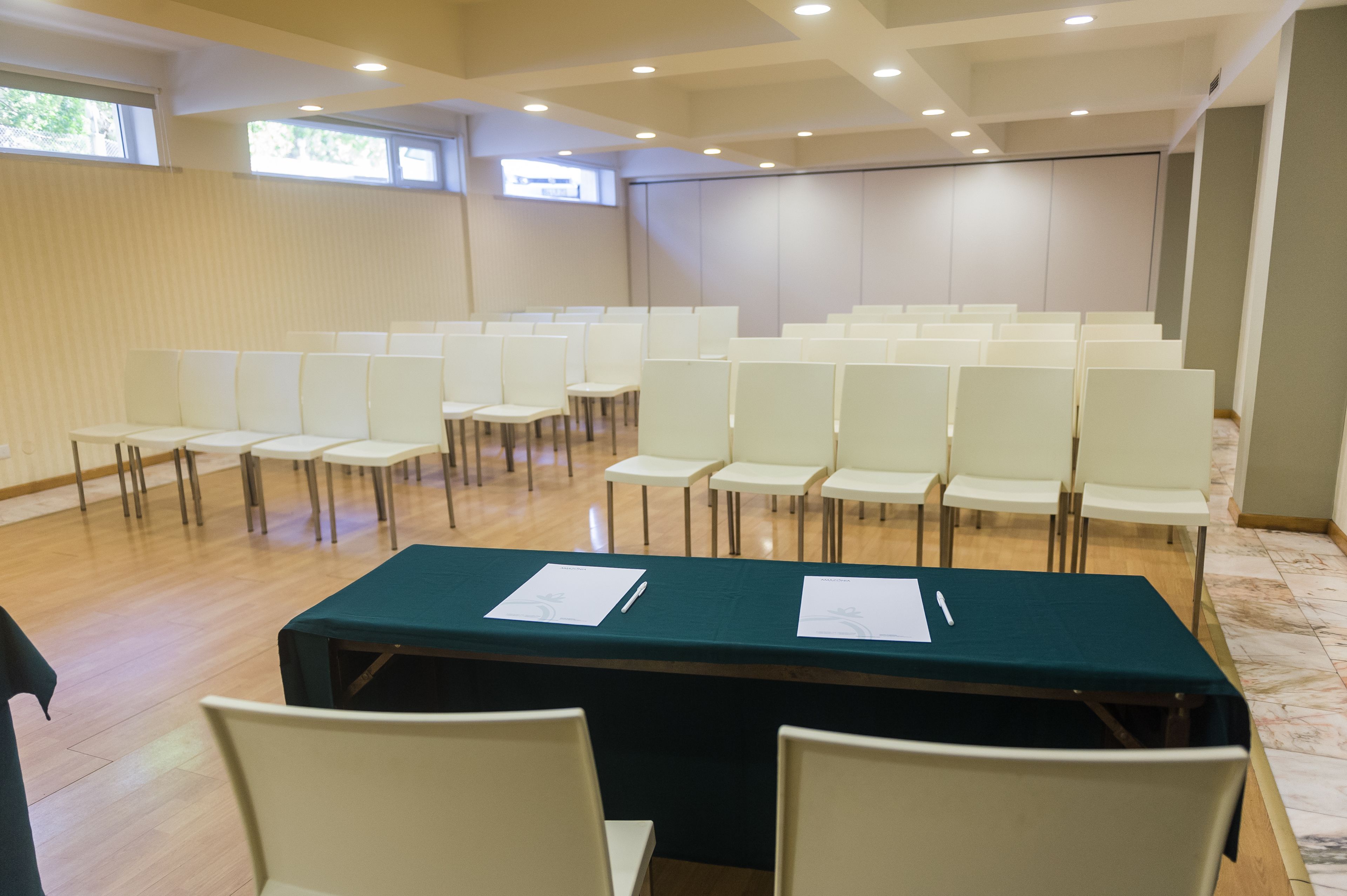 A conference room with rows of white chairs, a long table with papers, and a projector screen.