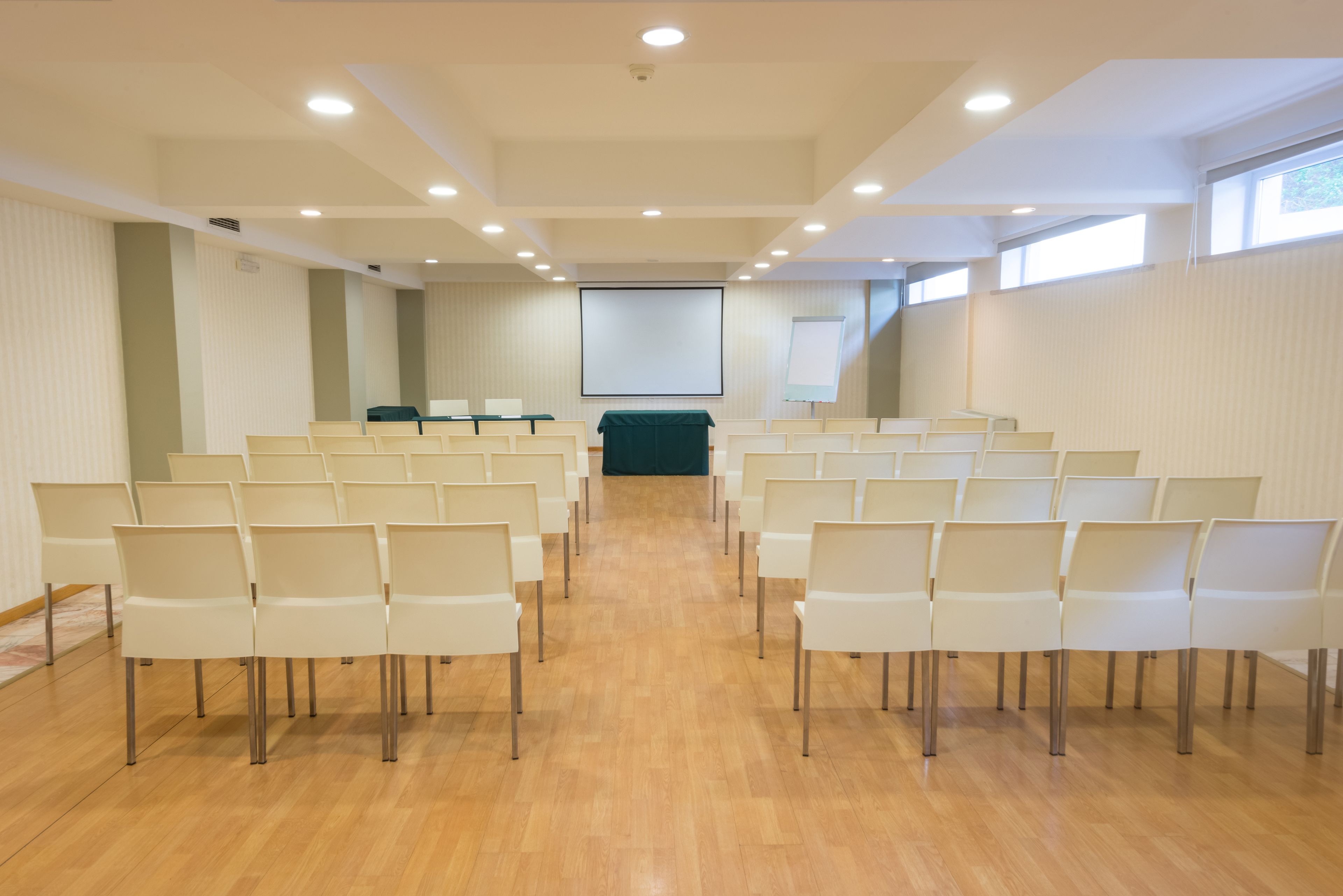 Una sala conferenze con file di sedie bianche, uno schermo per proiettore e uno schermo per presentazioni.