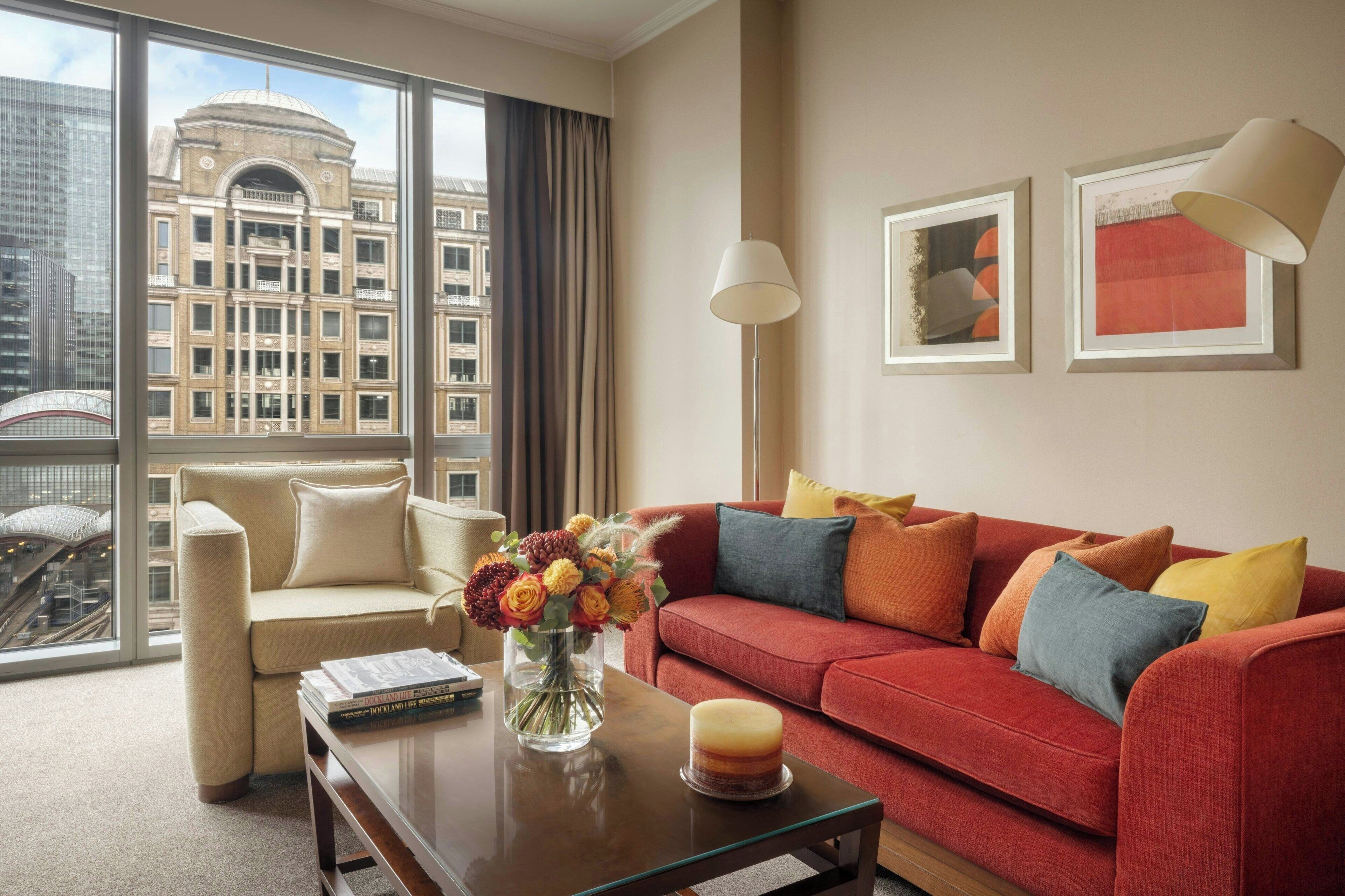 Een moderne hotelkamer met een rode bank, een beige fauteuil en een salontafel met een bloemenvaas. Een groot raam biedt uitzicht op een stadsbeeld.
