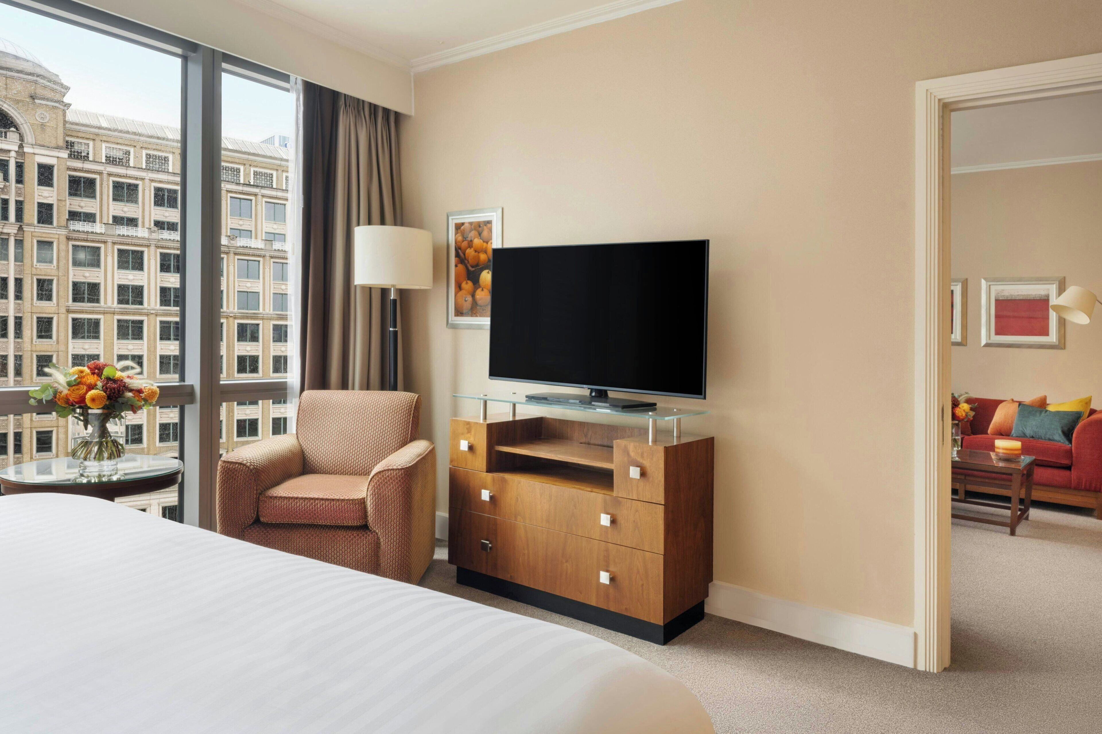 Habitación de hotel con una cama grande, un aparador de madera con un televisor de pantalla plana, una silla, un florero con flores y vista a un edificio desde la ventana.