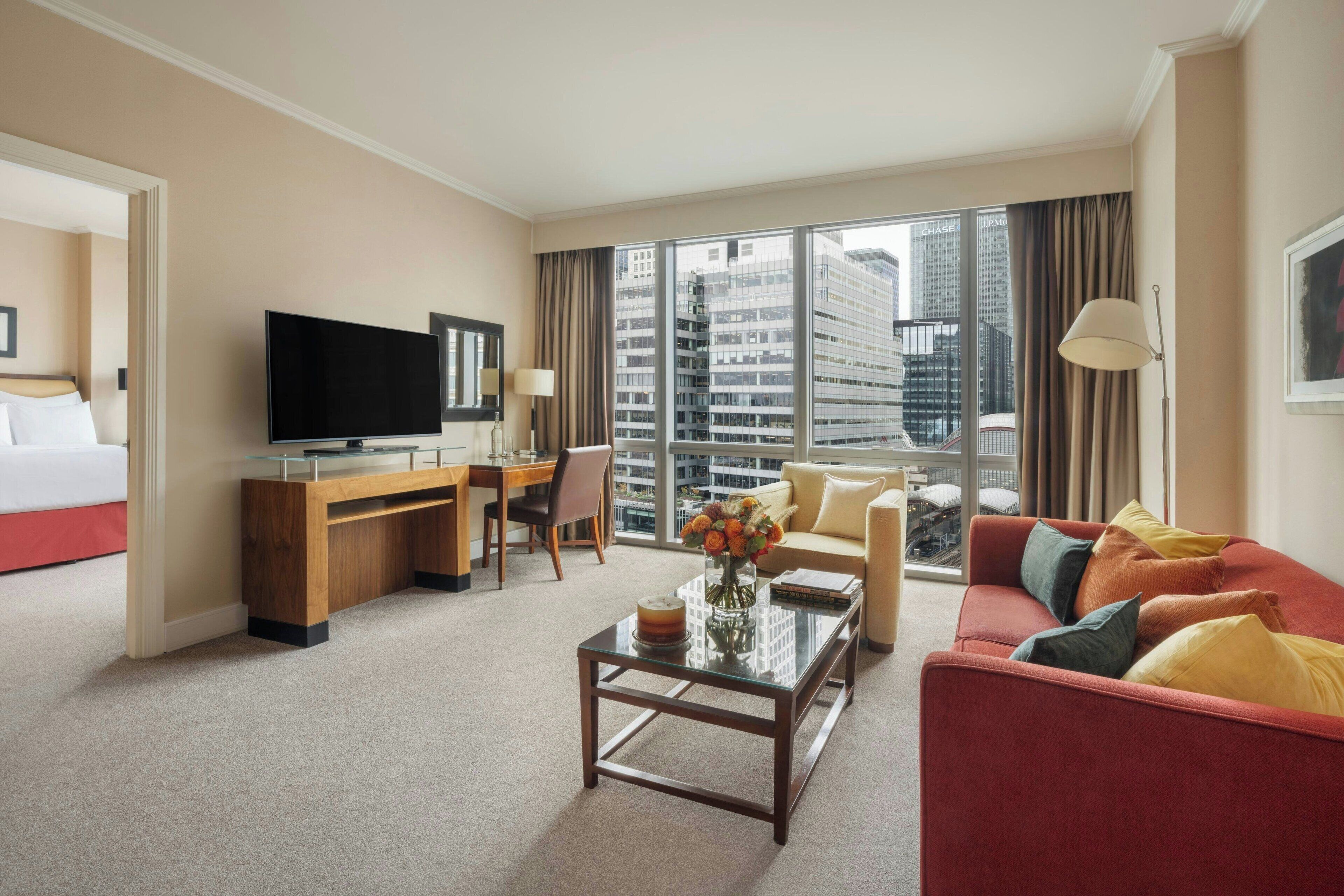 A hotel room with a bed, a desk with a TV, a coffee table with flowers, a red sofa, and a large window with a city view.