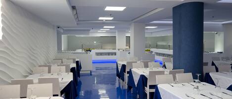 Un comedor moderno con mesas blancas, manteles azules y una barra iluminada en azul.