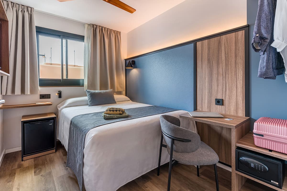 Camera d'albergo compatta con un letto, una scrivania, una sedia, un piccolo frigorifero e una valigia.