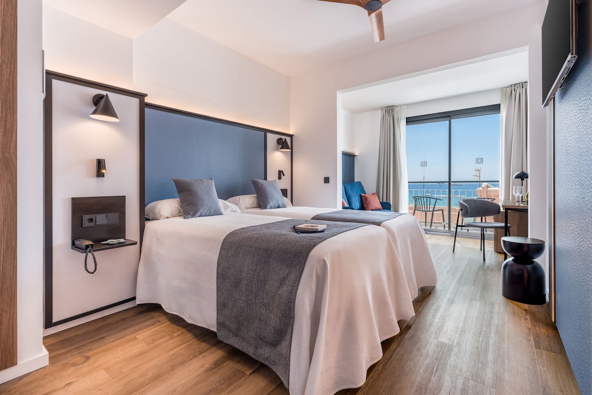 Camera d'albergo con due letti, una scrivania, una sedia, un balcone con vista e una ventola a soffitto.