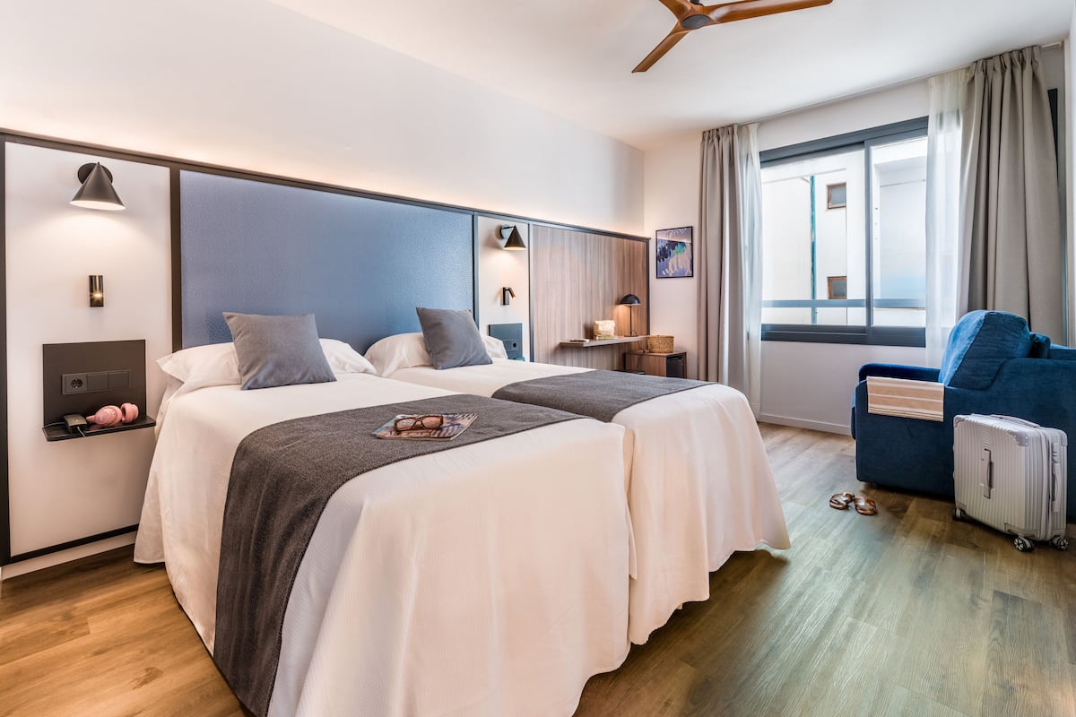 Una camera d'albergo con due letti, una scrivania, una sedia, una valigia e una finestra con tende.