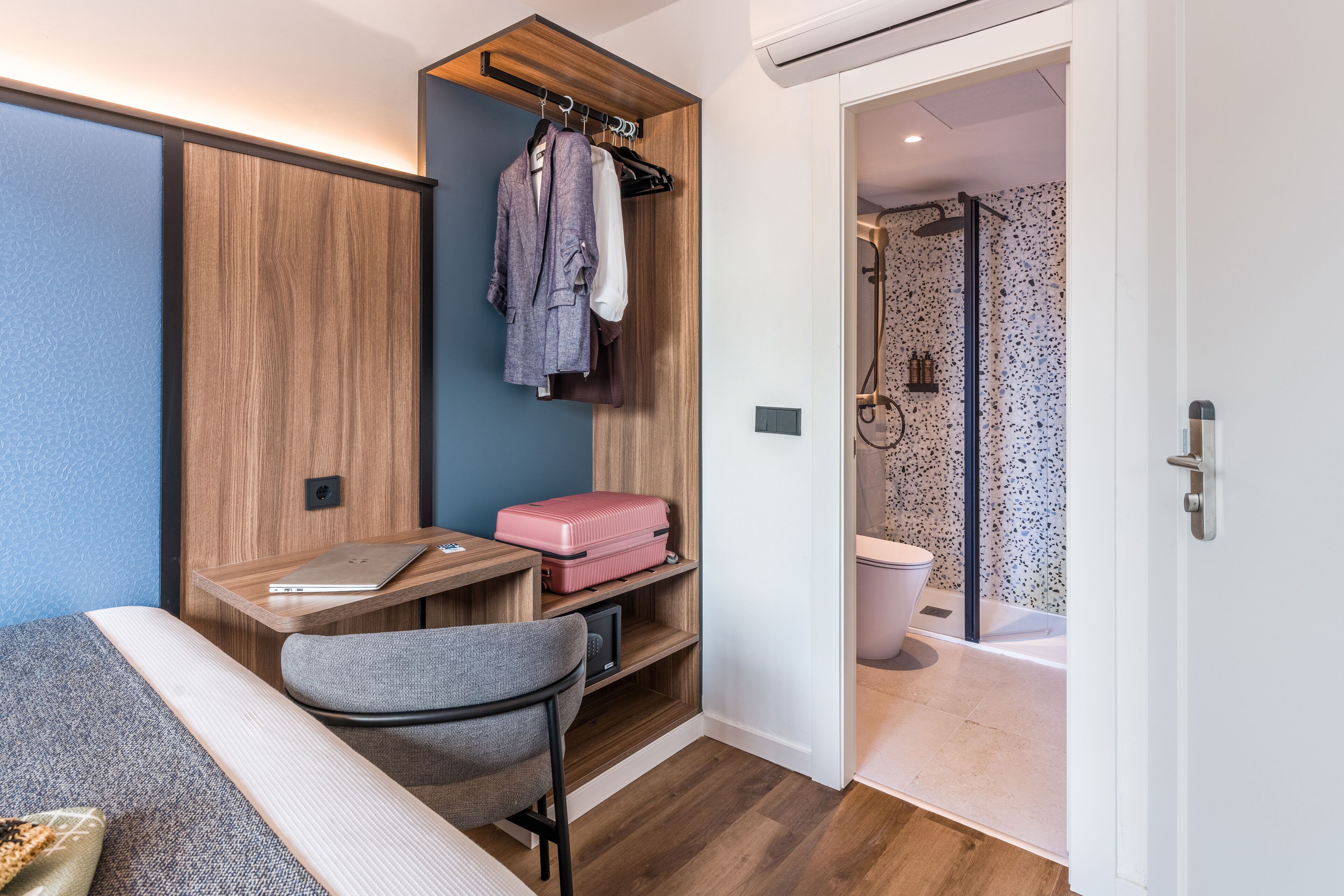 Ein kompakter Raum mit Schreibtisch, Stuhl und einer kleinen Sitzecke. Ein Kleiderschrank mit Kleidungsstücken und ein pinkfarbener Koffer sind sichtbar. Ein Duschbereich ist durch eine geöffnete Tür zu sehen.