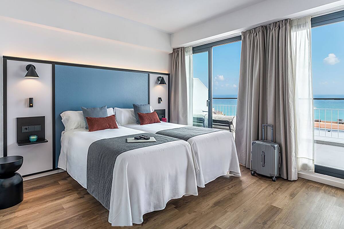 Camera d'albergo con un letto grande, una scrivania, una sedia, una valigia e un balcone vista mare.