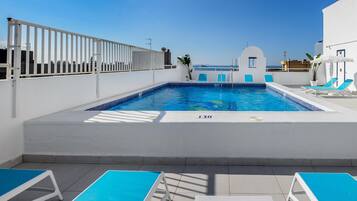 A rooftop swimming pool with a clear view of the sea, surrounded by white walls and blue lounge chairs.