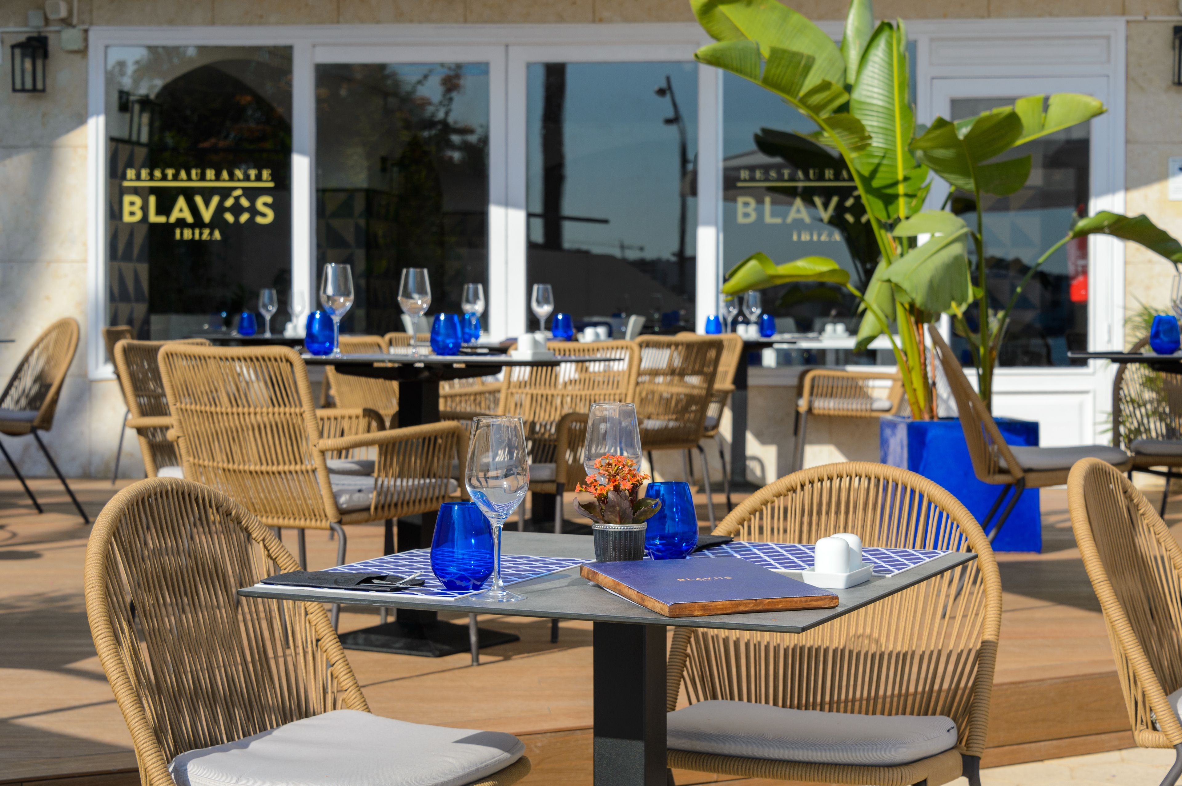 An outdoor dining area with wicker chairs and tables, blue glassware, and a potted plant.