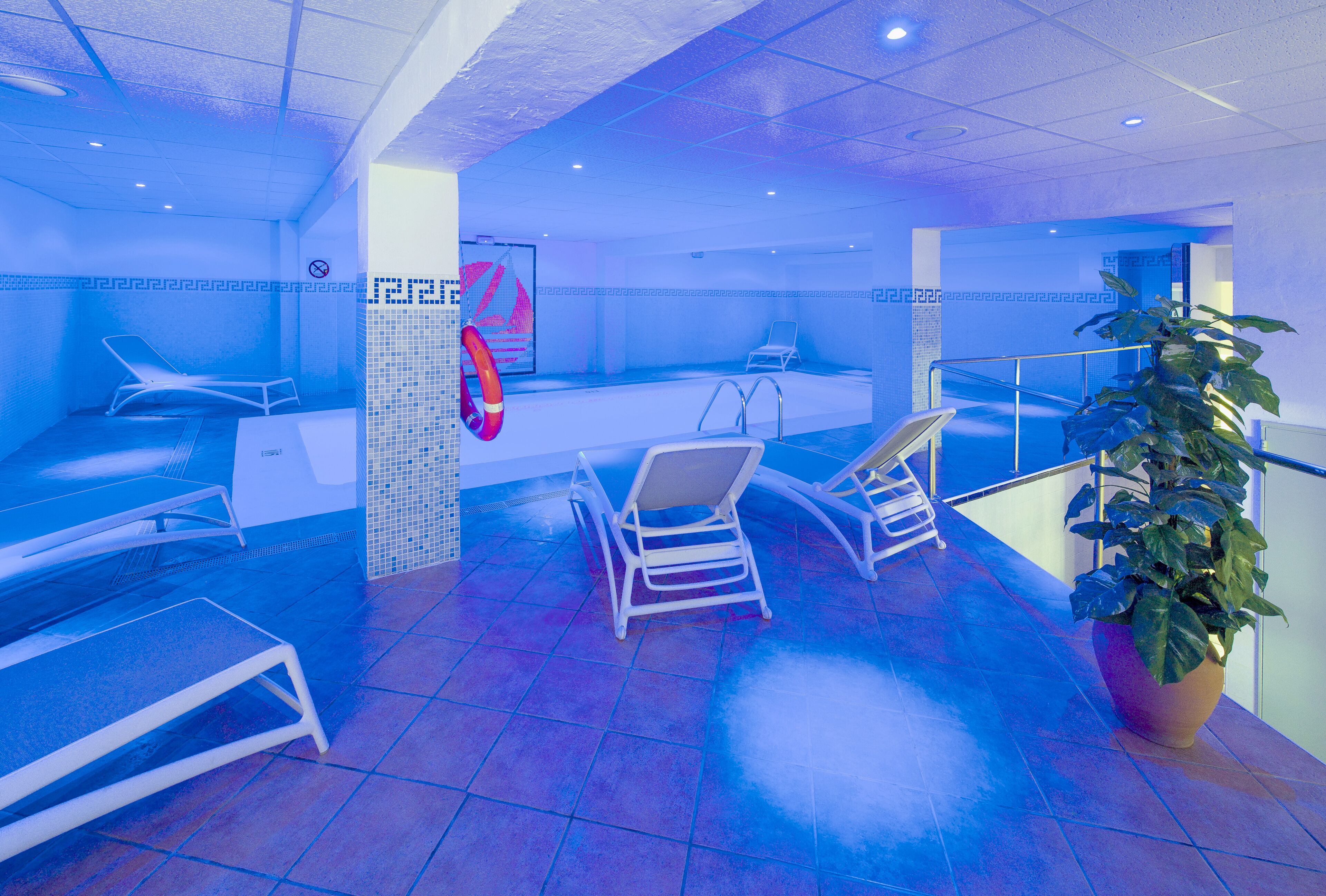 A blue-lit indoor swimming pool area with lounge chairs, a pool, and a potted plant.