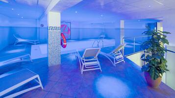 A blue-lit indoor swimming pool area with lounge chairs, a pool, and a potted plant.