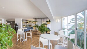 Un interior moderno con mobiliario blanco, una zona de cocina y amplias ventanas con vista a palmeras.