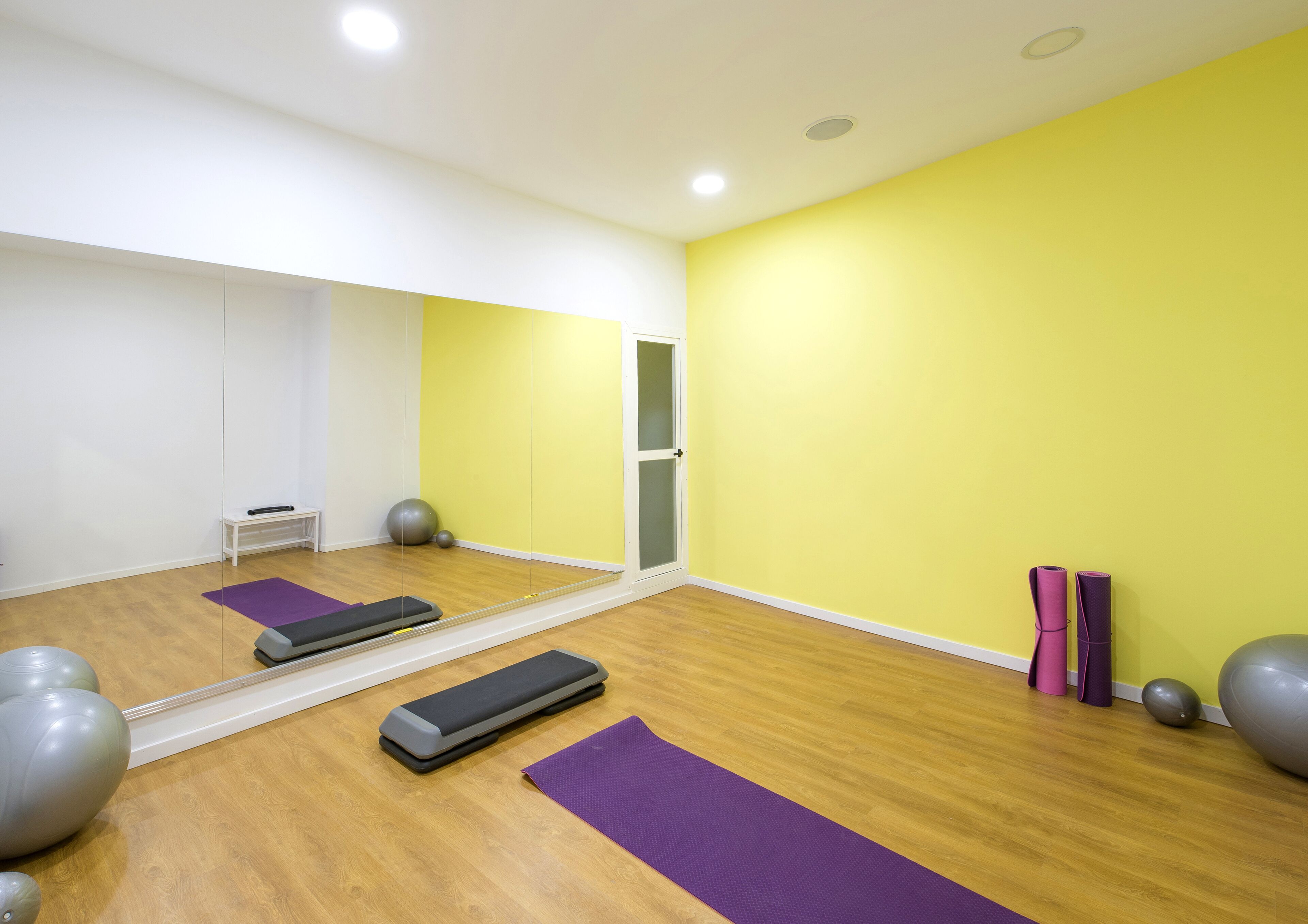 Een kamer met een grote spiegel, houten vloeren en fitnessapparatuur, waaronder een stepapparaat, yogamatten en fitnessballen.