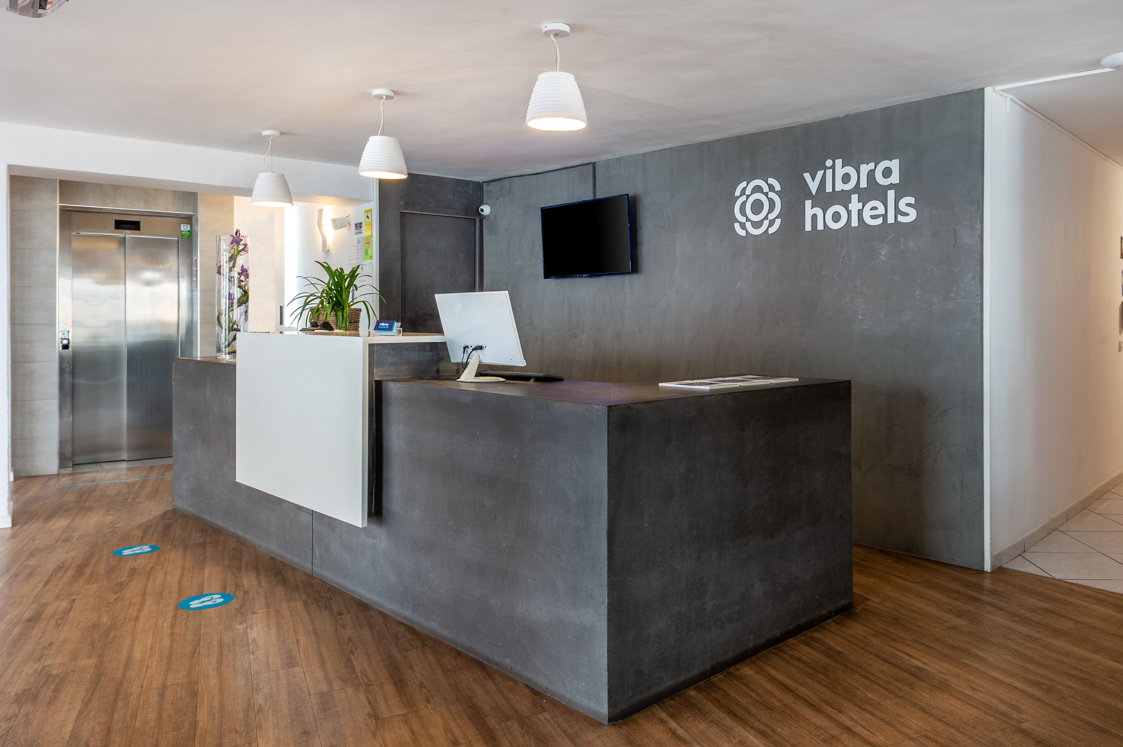 Een receptiebalie met een flatscreen-televisie, het logo van 'Vibra Hotels' en een modern interieur, met een grijs gebied voor de receptie en houten vloeren.