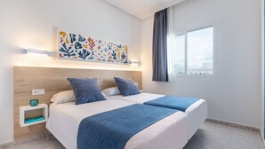 Habitación de hotel con una cama grande, dos almohadas, una manta azul, una obra de arte en la pared, una mesita de noche con un florero y una ventana con cortinas.