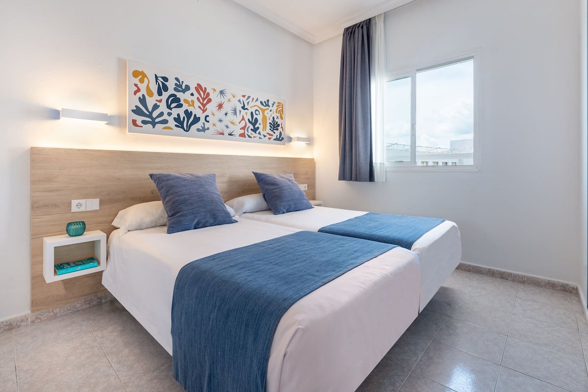 Une chambre d’hôtel avec un grand lit, deux oreillers, une couverture bleue, une œuvre d’art accrochée au mur, une table de chevet avec un vase et une fenêtre avec des rideaux.