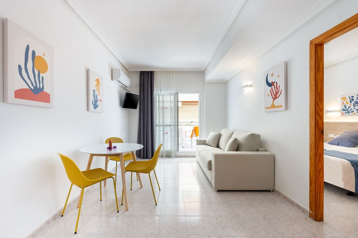 Une chambre d’hôtel moderne avec un canapé, une table à manger entourée de chaises jaunes et des œuvres d’art accrochées aux murs.