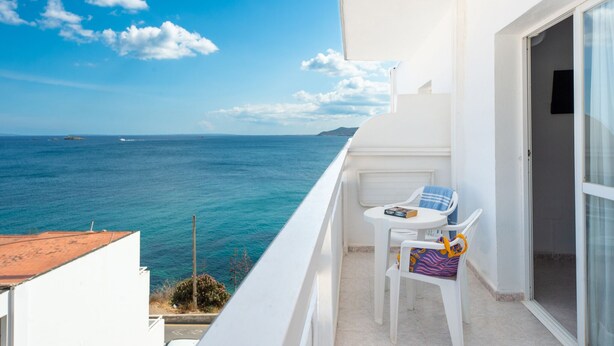 Un balcon donnant sur la mer, une table blanche avec une chaise, et une porte menant à un espace intérieur.