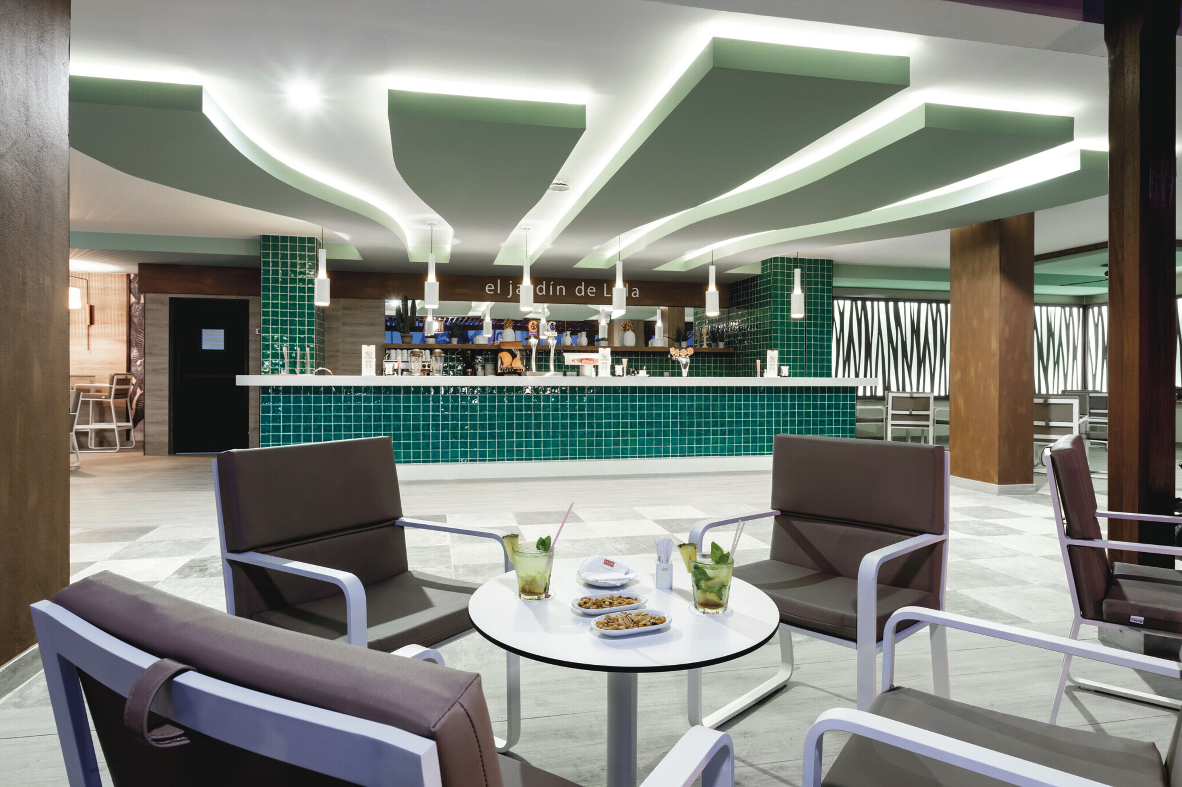 A modern bar area with a green-tiled counter, bar stools, and a round table with drinks and snacks.