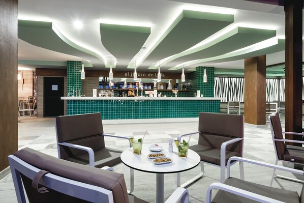 Zona de bar moderna con mostrador de azulejos verdes, taburetes y una mesa redonda con bebidas y aperitivos.