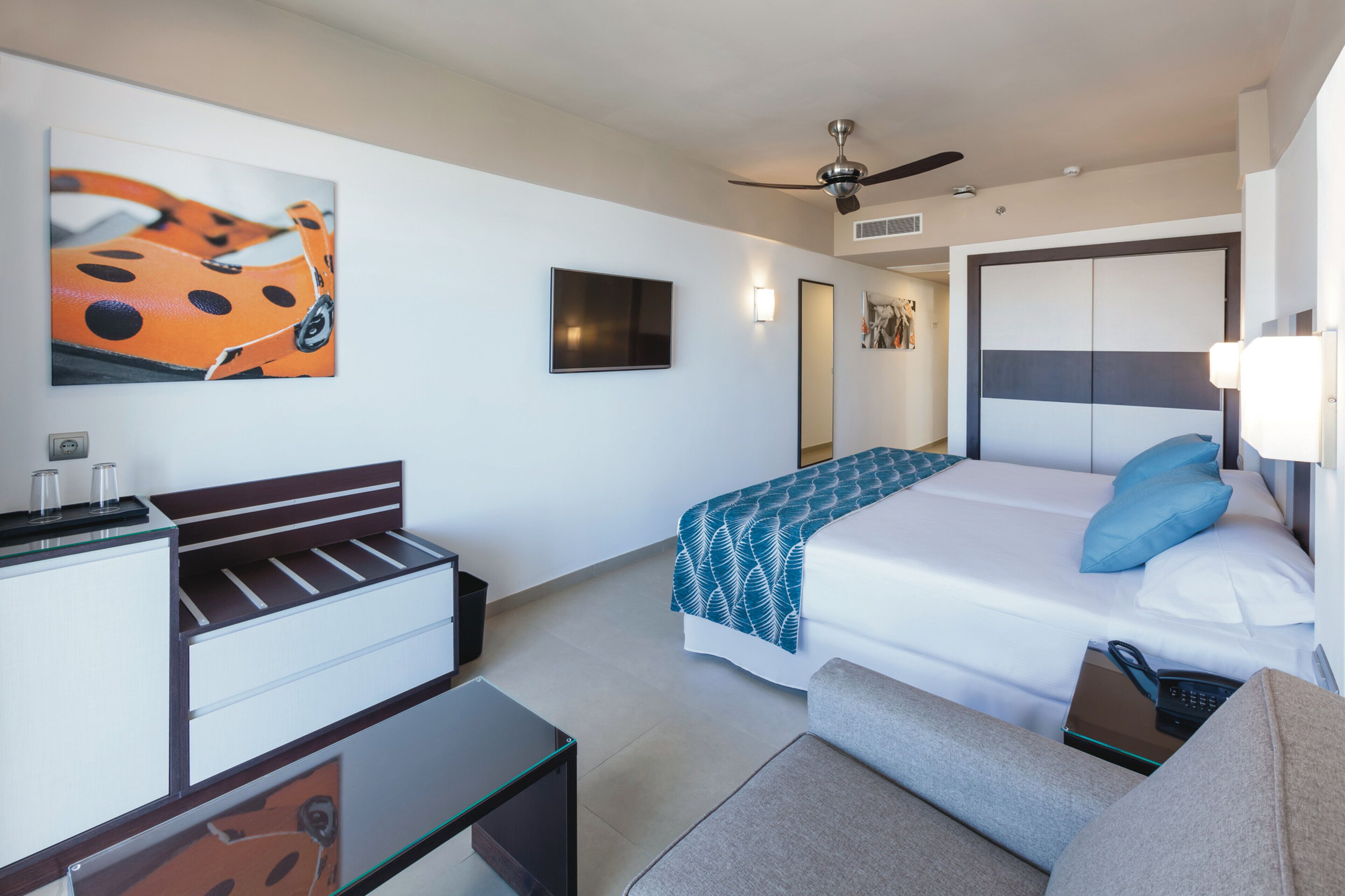 Een hotelkamer met een bed, een bureau, een stoel, een plafondventilator en een aan de muur bevestigde televisie.