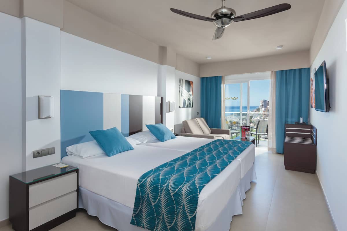 Quarto de hotel com cama grande, escrivaninha, cadeira e varanda com vista para a cidade.