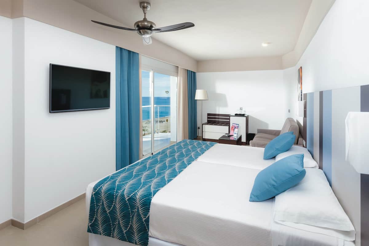 Quarto de hotel com cama, televisão, escrivaninha e vista para o oceano.