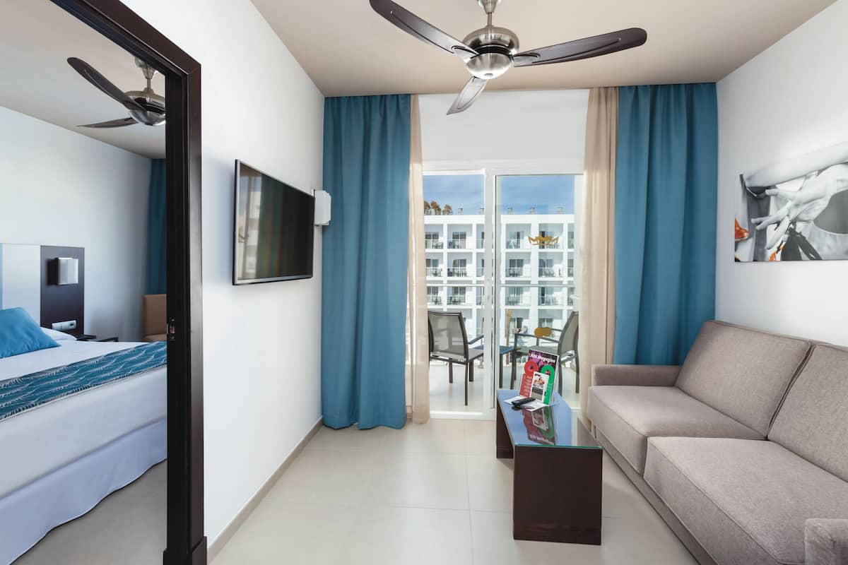 Quarto de hotel com cama, sofá, televisão, ventilador de teto e varanda com vista para edifícios.