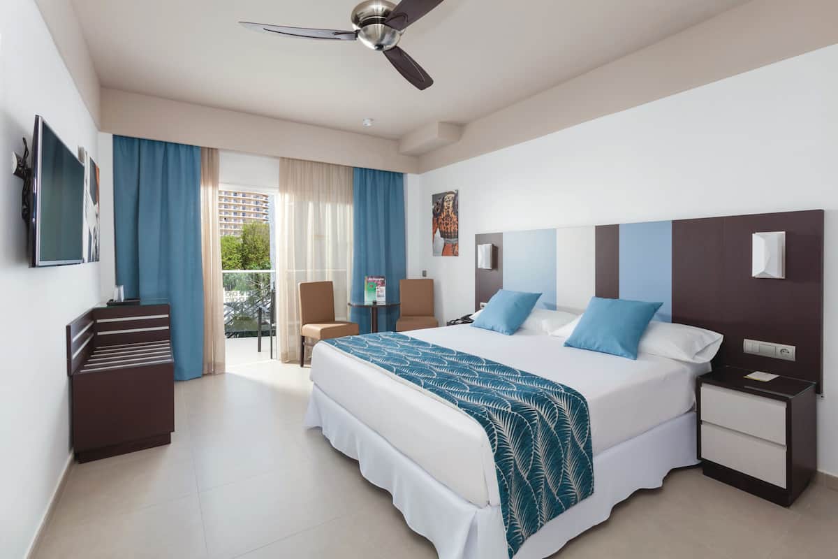 Quarto de hotel com cama, escrivaninha, cadeira, televisão e varanda com cortinas.