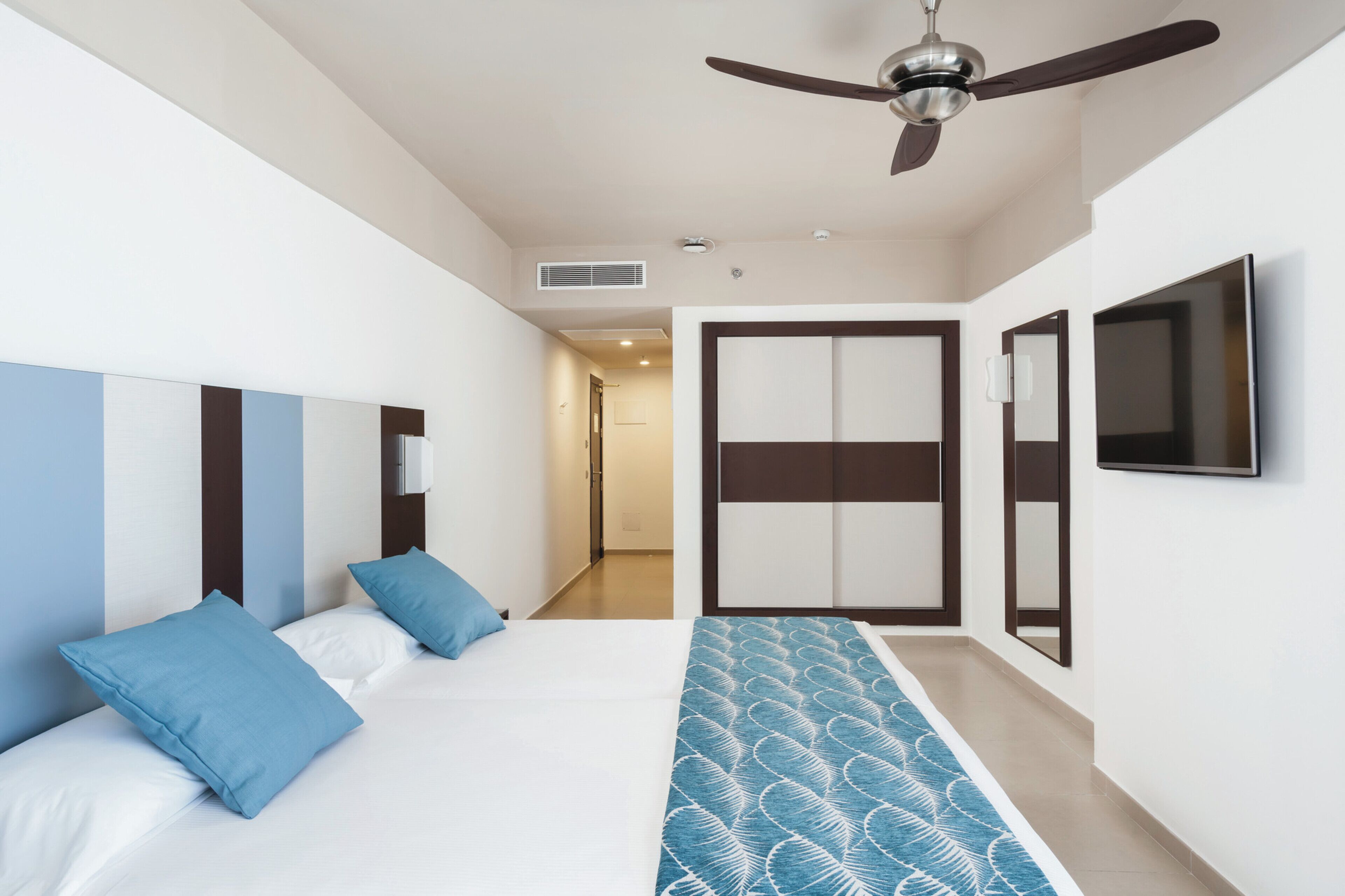 Habitación de hotel con cama, ventilador de techo, televisor y puerta que da a un pasillo.