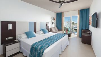 A hotel room with two beds, a ceiling fan, a TV, a desk, and a balcony with a view of the city.