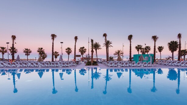 A poolside area with lounge chairs and palm trees, reflecting in the water. - Hotel Riu Costa del Sol - All Inclusive (Torremolinos)