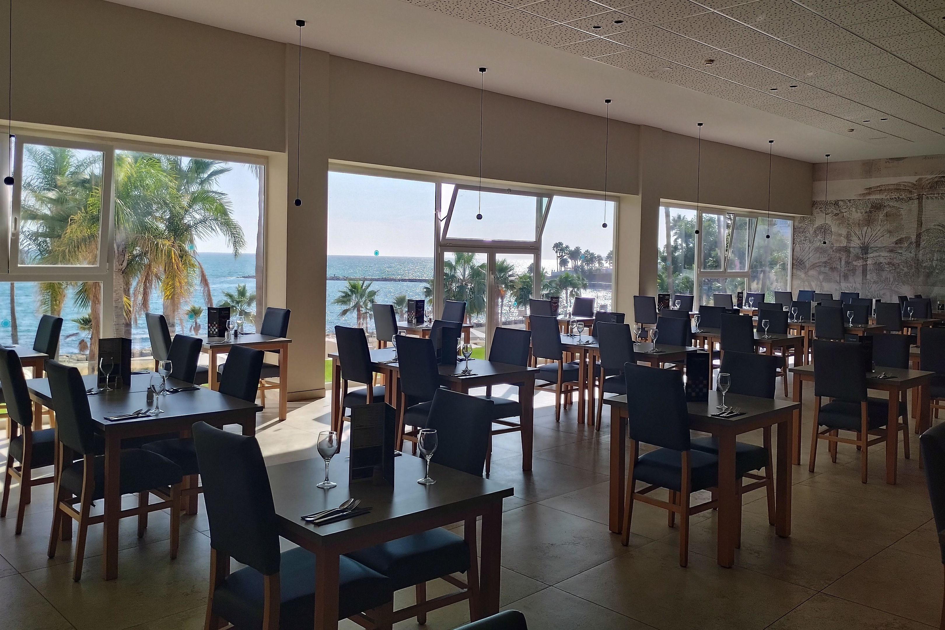 Un comedor con mesas y sillas, amplias ventanas con vista al mar y palmeras, y lámparas colgantes.