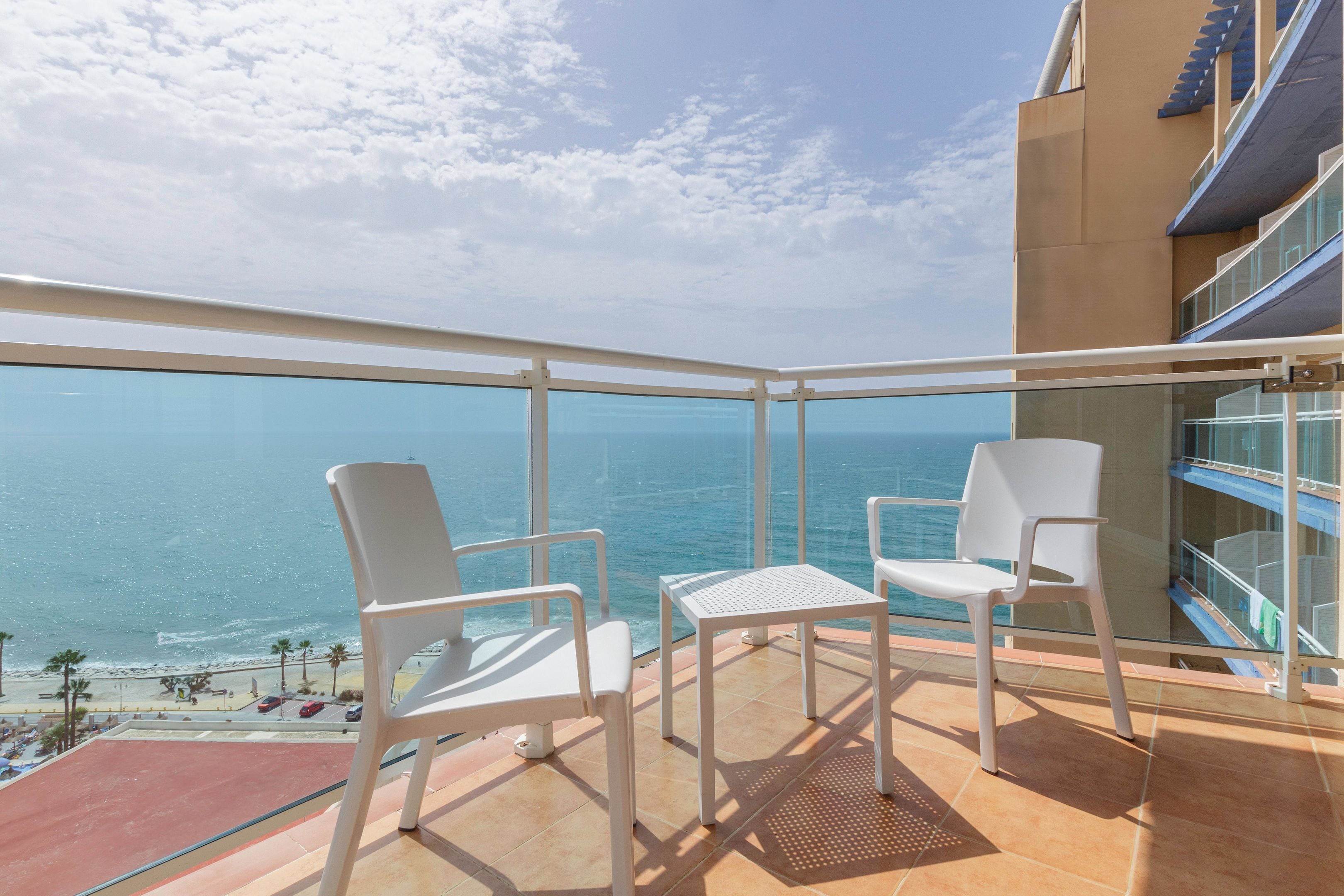 Un balcon avec deux chaises blanches et une petite table, donnant sur une plage et l’océan.