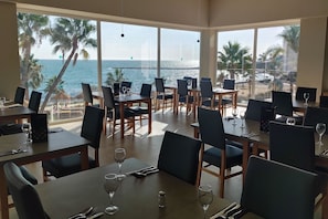 Um restaurante com mesas e cadeiras, grandes janelas com vista para a praia e oceano.
