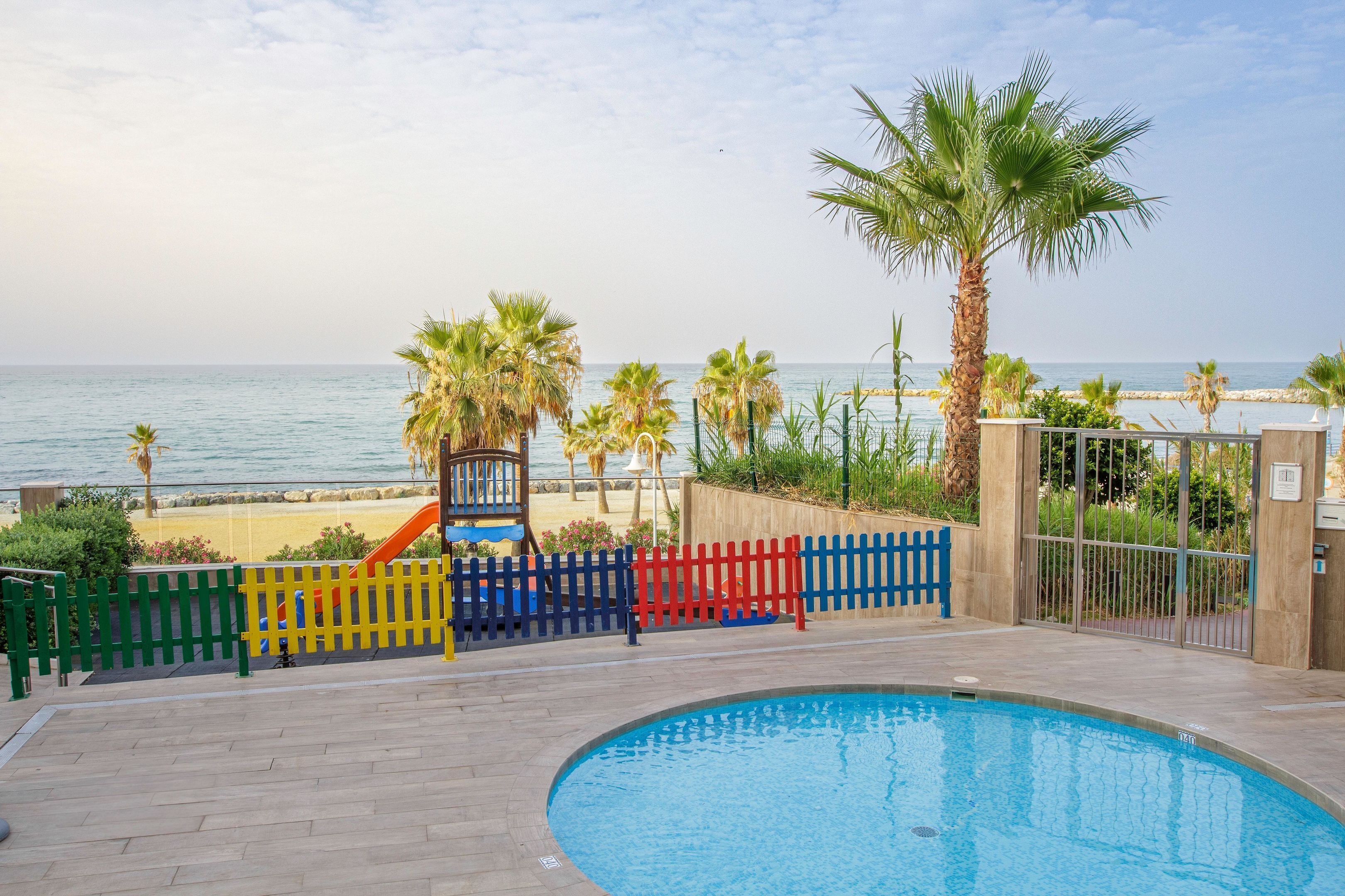 Piscina con vista al mar, rodeada de palmeras y una valla colorida.