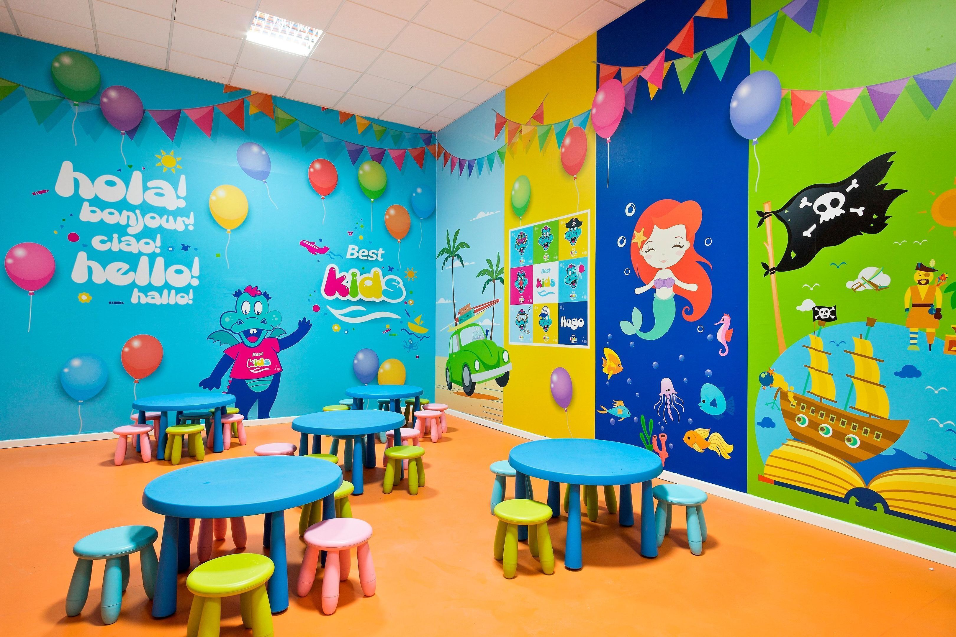 Un salón infantil lleno de color, con mesas y taburetes, decorado con globos y un mural de una sirena.