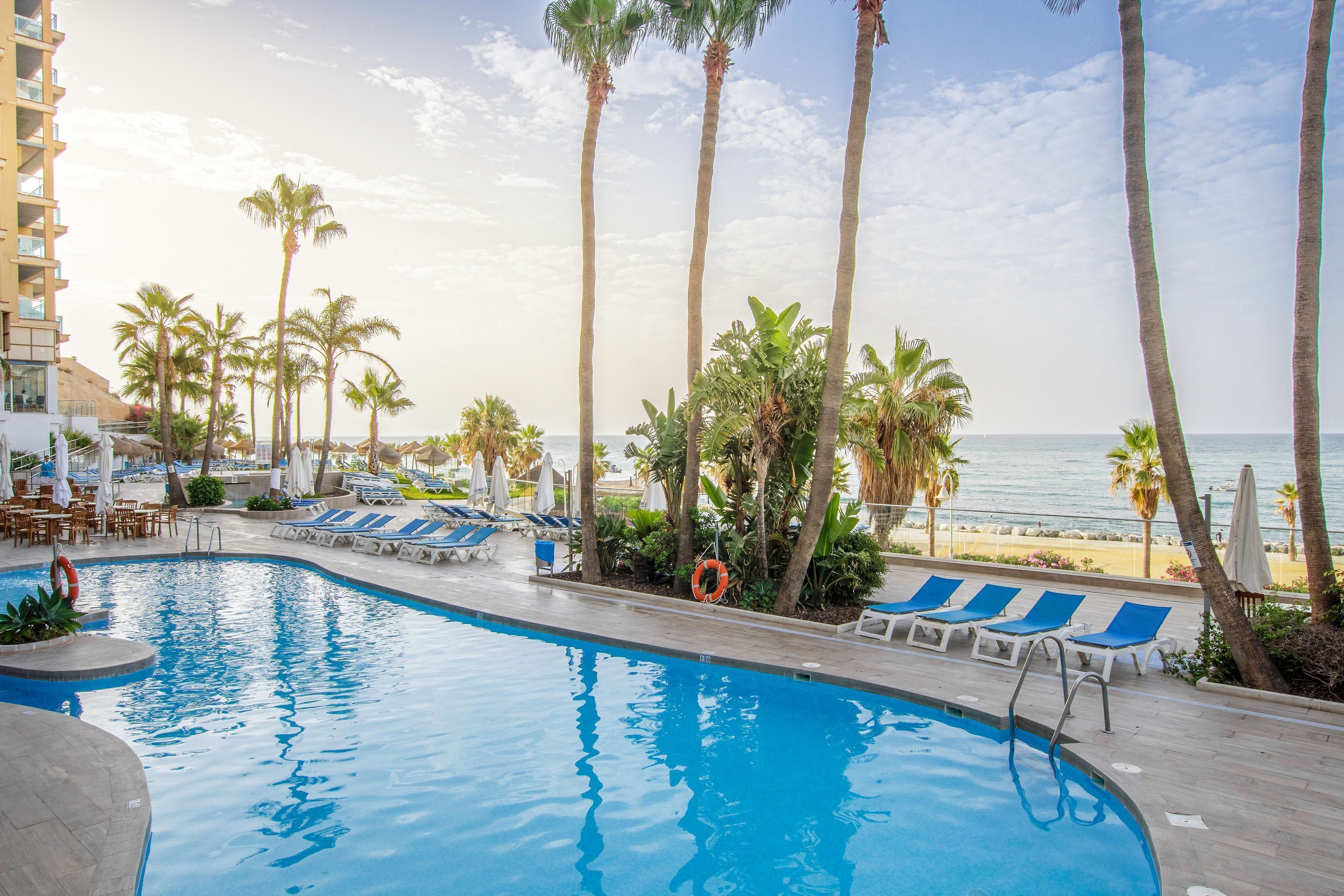 A swimming pool with lounge chairs and palm trees, overlooking a beach.