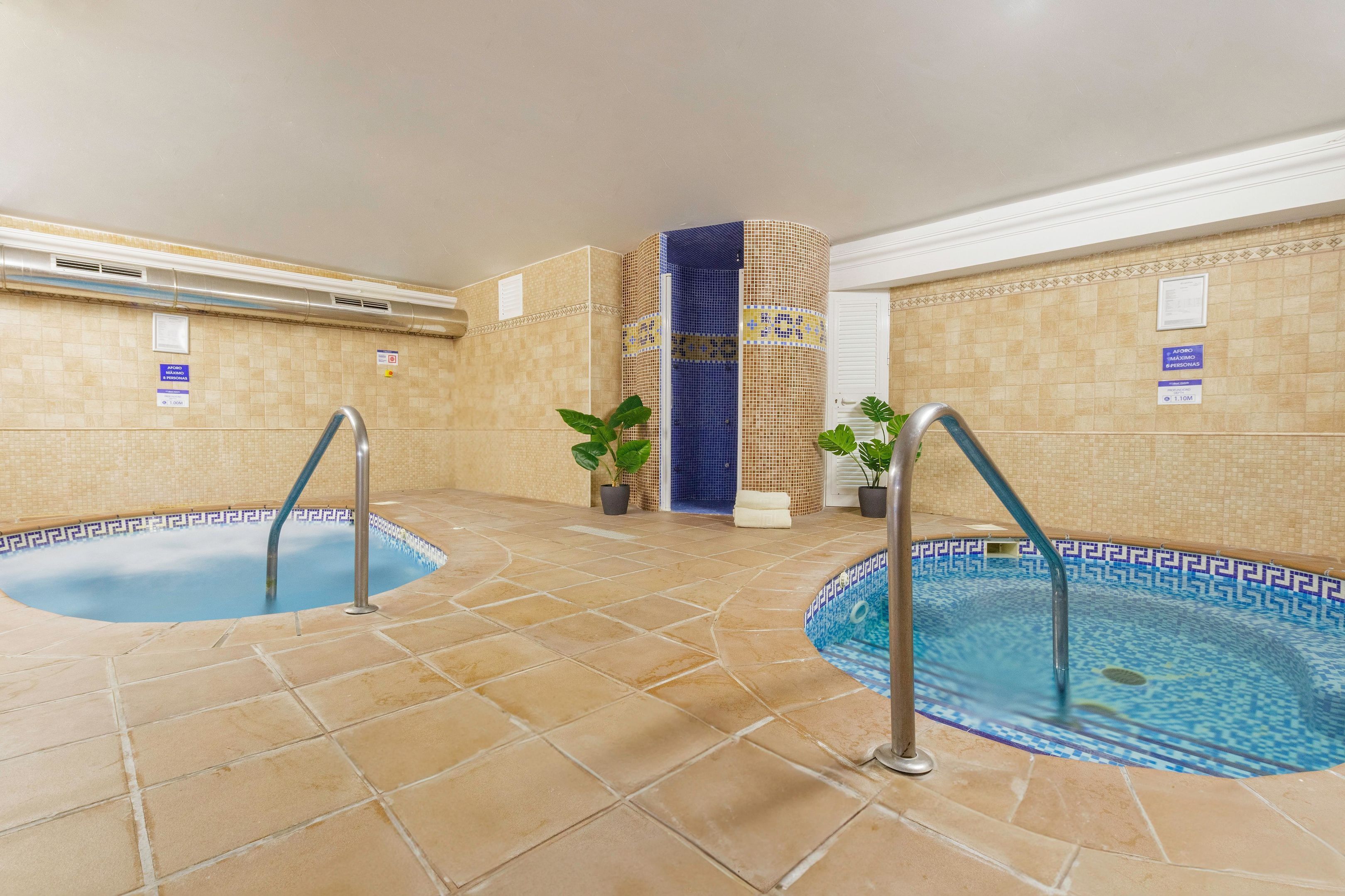 Zona de piscina de hotel con dos piscinas, paredes de azulejos y plantas en macetas.