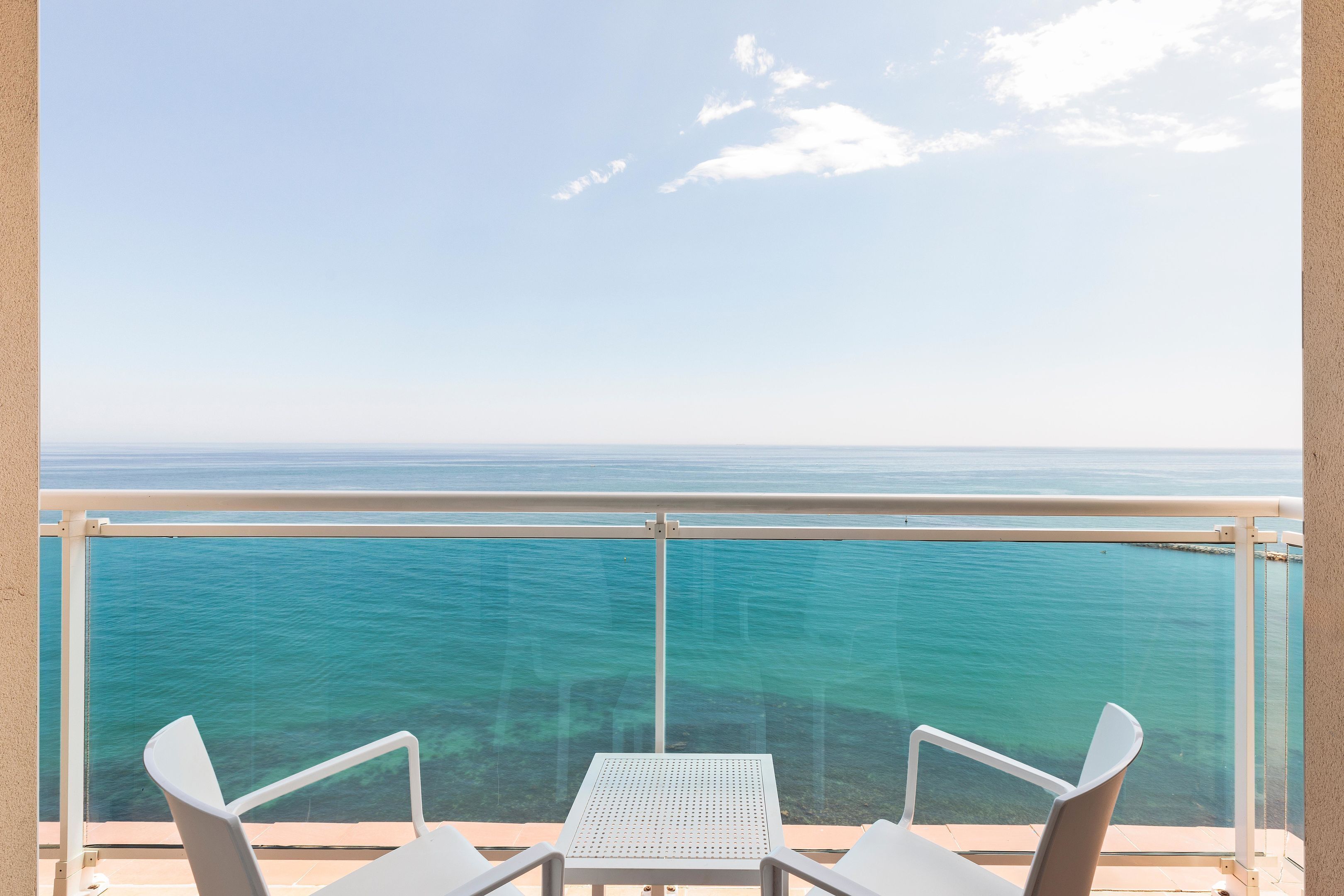 A balcony with a clear view of the ocean, two white chairs, and a small table.