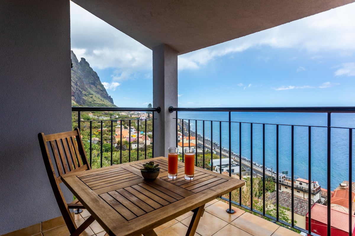 A wooden table with two glasses of red drink and a small plant on a balcony overlooking a coastal view with a mountain in the background.