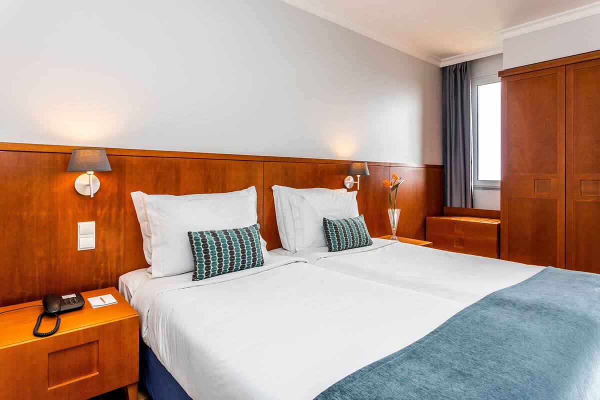 A hotel room with a wooden headboard, two beds with white bedding and patterned pillows, a bedside table with a lamp, a telephone, and a window with curtains.